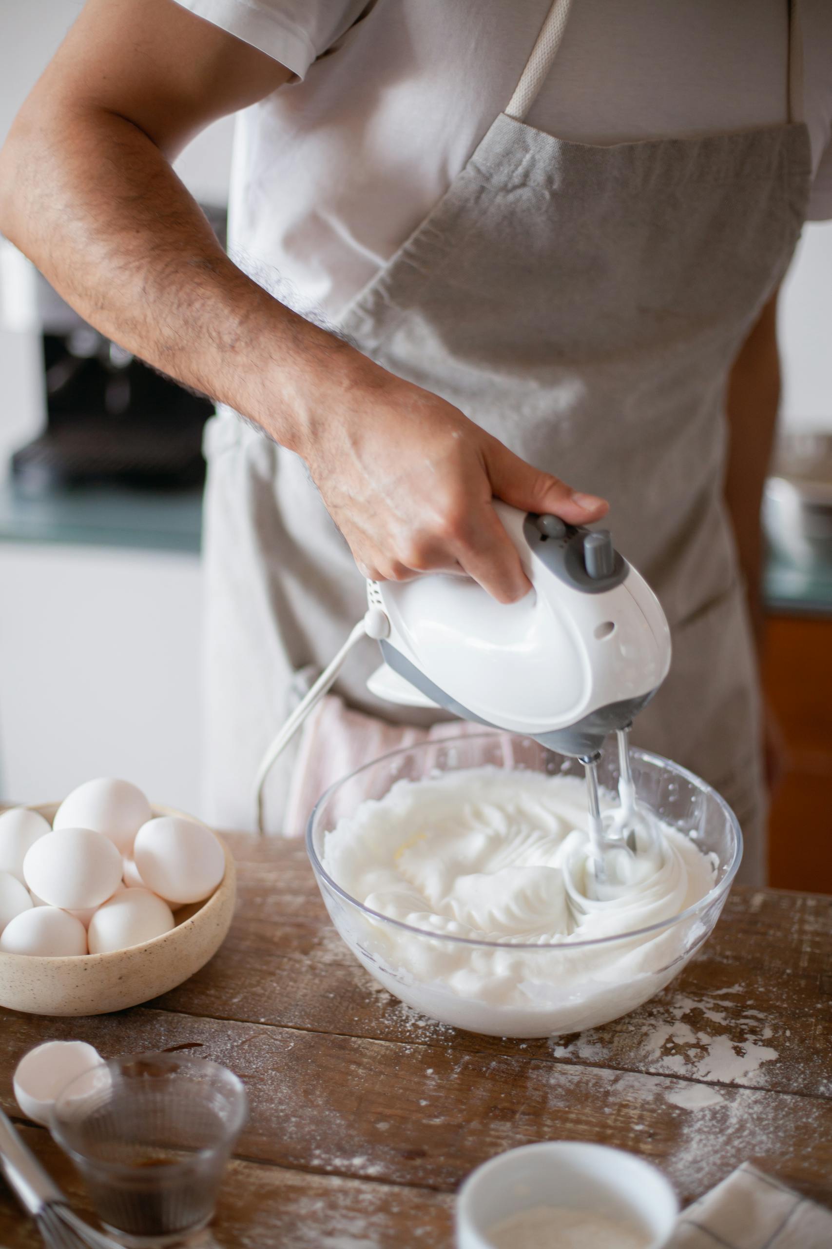 Male baker using hand mixer to whip cream in a glass bowl with eggs nearby.